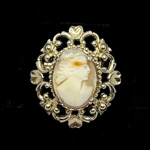 May include: Oval cameo brooch featuring a profile of a woman in cream against a light background. The cameo is set in a gold-toned filigree frame with floral and heart-shaped accents. The brooch is set against a black background.