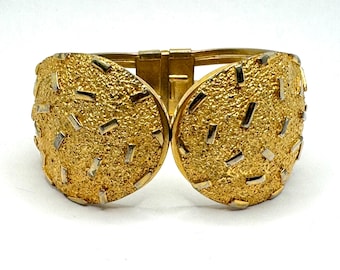 May include: A gold-toned hinged bracelet with a textured surface and rectangular silver accents. The bracelet has a clasp closure and a wide, rounded design. The jewelry piece is set against a white background.