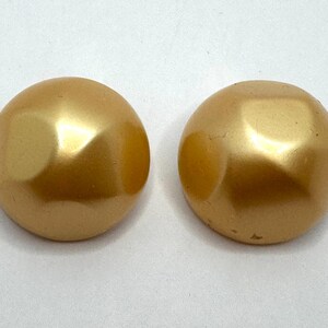 May include: Two gold-colored, round, faceted buttons. The buttons have a shiny, metallic finish and a slightly domed shape. The facets create a play of light and shadow on the surface.