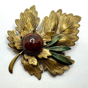 May include: A vintage brooch in the shape of a leaf. The brooch features a large, round, reddish-brown stone at the center, surrounded by gold-toned metal leaves. Green leaves are also present, adding a pop of color. The brooch is a unique accessory.