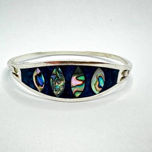 May include: A silver bangle bracelet with four oval-shaped abalone shell inlays set against a dark blue background. The iridescent shells display a range of colors, including green, blue, and pink. The bracelet is a piece of jewelry.