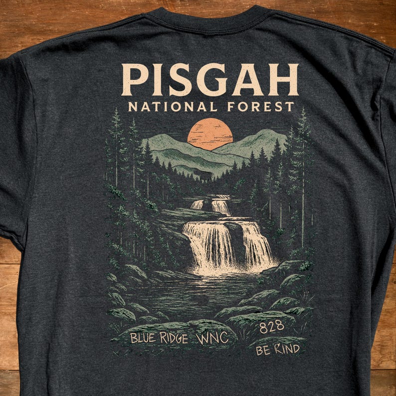 Może przedstawiać: Ciemnoszara koszulka z grafiką g&oacute;rskiego krajobrazu, wodospadu i zachodu słońca. Tekst "PISGAH NATIONAL FOREST" znajduje się na g&oacute;rze, z "BLUE RIDGE WNC" i "828 BE KIND" na dole.
