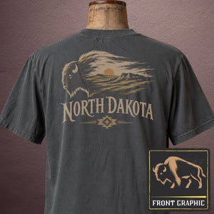 May include: A charcoal gray short-sleeve t-shirt featuring a back graphic of a bison silhouette integrated with a landscape, a sun, and the text North Dakota. A smaller inset box shows a bison graphic with the text Front Graphic below it.