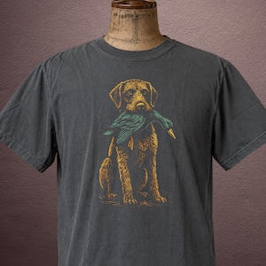 May include: A charcoal gray short-sleeve t-shirt featuring a graphic illustration of a golden-brown dog holding a teal and green duck in its mouth. The dog is depicted in a seated position with detailed line work, centered on the front of the shirt.