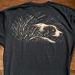 May include: Dark gray t-shirt featuring a detailed illustration of a hunting dog in tall grass. The dog is depicted in shades of brown and white, with a focused expression. The design is printed on the back of the shirt.