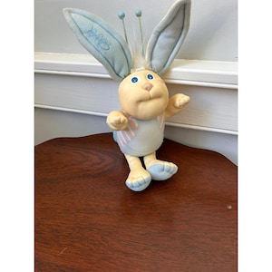 Puede incluir: Un peluche que representa una criatura de color amarillo claro con orejas largas de color azul claro, ojos azules y dos antenas con puntas azules. El juguete lleva un traje azul claro con rayas rosas y blancas en el pecho y tiene un mechón de pelo blanco.