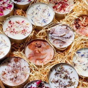 Könnte beinhalten: Nahaufnahme von mehreren kleinen, runden Kerzen in Metallbehältern. Das weiße Wachs ist mit getrockneten Blumen und Kräutern verziert. Die Kerzen sind auf einem Strohbett angeordnet, was einen natürlichen und rustikalen Eindruck vermittelt.