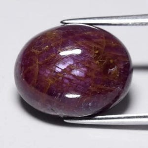 May include: An oval-shaped, cabochon-cut ruby gemstone with a deep, saturated magenta-red color. The stone features a smooth, polished surface with visible internal inclusions and a network of fine, golden-brown veining throughout the body.