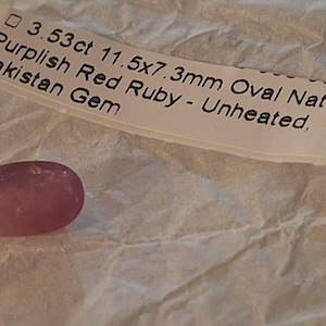 May include: A purplish red oval ruby gemstone with a faceted surface. A label next to the gem reads: 3.53ct 11.5x7.3mm Oval Natural Purplish Red Ruby - Unheated, Pakistan Gem. The gemstone is a deep pinkish-red colour with a translucent appearance.