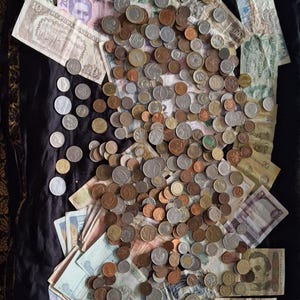 Op de afbeelding: Een verzameling internationale munten en bankbiljetten, waaronder Amerikaanse dollars en euro's. De munten variëren in grootte en kleur, met zilver- en kopertinten. Sommige bankbiljetten zitten in plastic hoesjes.