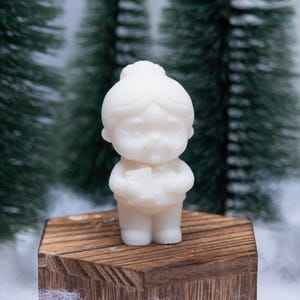 Może przedstawiać: Mała, kremowa figurka kobiety z upiętymi włosami, trzymającej książkę. Figurka stoi na drewnianym bloku o sześciokątnym wierzchu. Tło przedstawia miniaturowe drzewka iglaste i efekt śniegu.