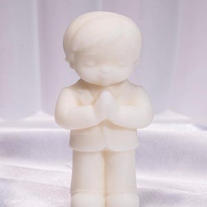 Può includere: Una piccola statuina color crema di un ragazzino in abito, con le mani giunte in preghiera. La figura ha una finitura liscia ed è in piedi. Lo sfondo è un morbido tessuto bianco.