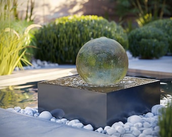 Könnte beinhalten: Ein Wasserbrunnen mit einer großen, durchscheinenden, kugelförmigen Glaskugel, die auf einem glatten, schwarzen, quadratischen Sockel zentriert ist. Wasser fließt über die Oberfläche der Kugel und erzeugt einen Welleneffekt beim Herunterlaufen.