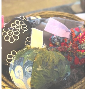 May include: A collection of fabric pumpkins in a woven basket. One pumpkin features a black fabric with white flower outlines, another has a green and blue patterned fabric with a cream stem, and a third displays a red floral print fabric.