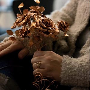 May include: A bouquet of copper roses with detailed petals and leaves. The flowers are held in a hand, with a ring on the finger. The copper has a warm, metallic sheen. The stems are bundled together.
