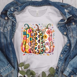 Peut inclure: Un t-shirt blanc avec un motif de trois citrouilles. La citrouille de gauche présente des pois colorés, celle du centre un motif léopard, et celle de droite un design floral. De petites étoiles et des feuilles entourent les citrouilles.