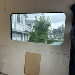May include: Interior view of a van with a window showing a residential neighborhood. The walls are light-colored wood panels with a rectangular cutout and a power outlet. The window has a black frame.