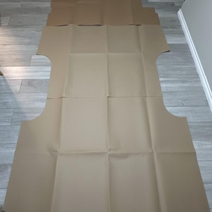 May include: A large, unfolded sheet of cardboard, possibly for packaging or a craft project, is laid out on a light grey wood-effect floor. The cardboard is light brown and folded into rectangular sections.