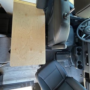 May include: A light-colored wooden table installed in a vehicle. The table is rectangular and appears to be made of plywood. The interior of the vehicle includes black leather seats, a steering wheel, and a gray floor.