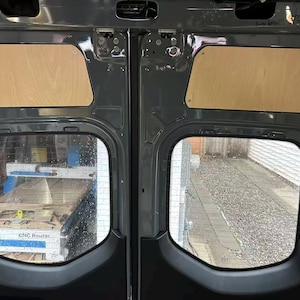 Puede incluir: Vista interior de las puertas traseras de una furgoneta, con dos ventanas y paneles de madera. Las puertas son grises, con una vista de una fresadora CNC visible a través de una ventana y un camino de grava a través de la otra. Los paneles de madera son rectangulares.