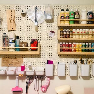 May include: A pegboard wall organizer with various art supplies. Includes paint bottles, brushes, containers, and shelves. The color palette is vibrant, with a mix of paints and tools for creative projects. The pegboard is white with black dots.