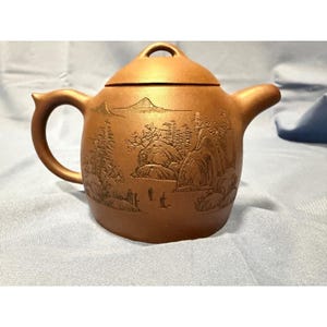 May include: A brown ceramic teapot with a lid and handle. The teapot has an engraved landscape design with mountains, trees, and figures. The spout and handle are curved. The teapot is likely used for brewing tea.