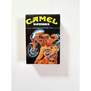 May include: A rectangular black box featuring the yellow text CAMEL SUPERBIKE at the top. The graphic depicts an orange camel wearing a racing suit with the text CAMEL PRO, standing next to an orange racing motorcycle with the number 1.
