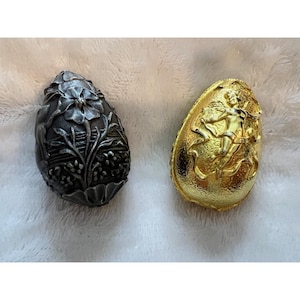 May include: Two decorative egg-shaped ornaments. One is dark gray with a floral design, the other is gold-colored with a relief of a figure with a bow and arrow. Both are on a soft, white surface.
