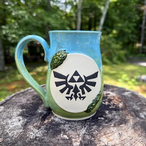 May include: A ceramic mug with a gradient blue and green glaze featuring a central white circular medallion. The medallion displays a black Triforce emblem with wings. Two textured, moss-like green decorative accents flank the white circle.