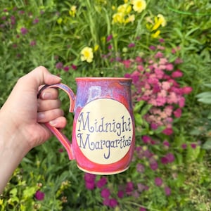 May include: A ceramic mug with a glossy glaze in shades of pink, purple, and blue. The mug features a central cream-colored circular emblem with the text Midnight Margaritas written in a dark purple, stylized cursive font on the front surface.