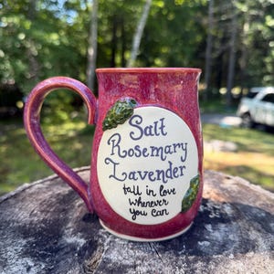 May include: A ceramic mug with a glossy magenta and purple glaze featuring a central cream-colored oval panel. The text reads Salt, Rosemary, Lavender, fall in love whenever you can. Two small green sculpted leaf accents flank the text panel.
