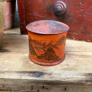 May include: A round, orange tin with a lid, decorated with a brown illustration of a person, mountains, and trees. The tin is placed on a wooden surface.