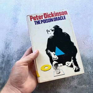 May include: A vintage paperback book titled "The Poison Oracle" by Peter Dickinson. The cover features a black silhouette of a monkey with a blue triangle and yellow circle on a white background. The book is held by a hand.
