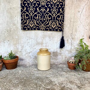 May include: A vintage stoneware jar with a cream body and a tan rim. Small terracotta plant pots with succulents and greenery are arranged around the jar. A navy blue and gold patterned textile hangs in the background. The jar is in a rustic setting.
