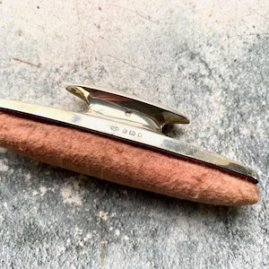 May include: A silver nail buffer with a pink felt pad. The buffer has a decorative design on the top and the inscription "925" on the side.