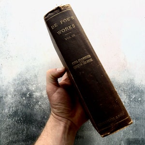 May include: A vintage brown leather-bound book titled "DE FOE'S WORKS" with the volume number "VOL. III" and the title "MOLL FLANDERS and HISTORY OF THE DEVIL" printed on the spine. The book is held in a hand against a white background.