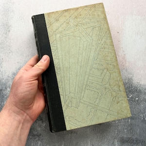 May include: A vintage hardback book with a black spine and a beige cover featuring a line drawing of a tall building with surrounding streets and trees.
