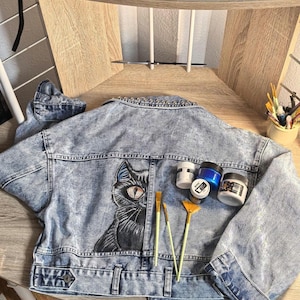 Könnte beinhalten: Eine hellblaue, säuregewaschene Jeansjacke mit einem handgemalten schwarzen Katzenmotiv und einem markanten Auge auf der Rückseite. Die Jacke ist am Kragen mit silbernen Metallnieten verziert und mit drei Pinseln sowie vier kleinen Acrylfarbtöpfen belegt.