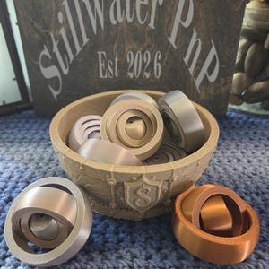 May include: A beige plastic bowl with a shield emblem containing the letter S, filled with several spherical fidget toys in metallic silver, gold, and copper finishes. Two additional spherical fidget toys sit in front of the bowl.