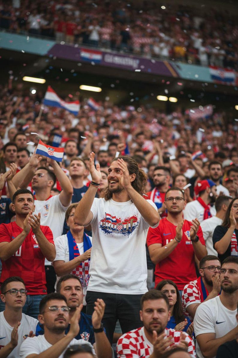 Pu&ograve; includere: Una folla in uno stadio, molti indossano camicie a scacchi rosse e bianche o rosse e sventolano bandiere croate. Un uomo al centro indossa una maglietta bianca con un disegno della bandiera croata e applaude.