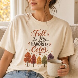 May include: A cream-colored short-sleeve t-shirt featuring the text "Fall Is My Favorite Color" in brown and orange script and block lettering, accompanied by a graphic of five stylized autumn trees in shades of orange, red, yellow, and purple.