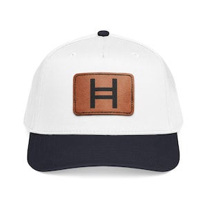May include: White baseball cap with a navy blue brim and a brown leather patch. The patch features a black letter "H" with a horizontal bar across the middle. The cap has a black button on top.