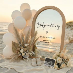 Op de afbeelding: Een strandscène met een houten spiegel met de tekst "Baby on the way". De scène bevat crèmekleurige ballonnen, pampasgras, bloemen, babyslofjes en een echo-afbeelding. De tekst bevat ook de achternaam van de baby en de verwachte aankomstmaand.