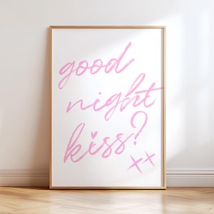 May include: A framed art print with the words "good night kiss?" in pink script. The artwork is displayed on a white wall and rests on a wooden floor. The frame is a light brown color.