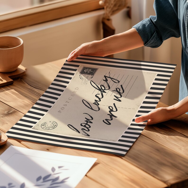 K&ouml;nnte beinhalten: Eine dekorative Postkarte mit dem Text "how lucky are we" in handschriftlichem Stil. Die Postkarte hat einen schwarz-wei&szlig; gestreiften Rand und eine Vintage-&Auml;sthetik. Das Bild befindet sich auf einer Holzoberfl&auml;che.