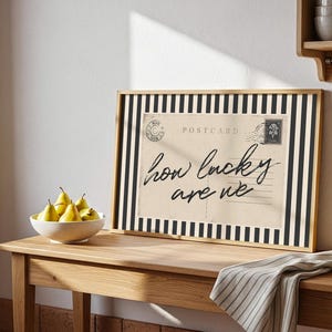 K&ouml;nnte beinhalten: Ein gerahmter Postkartendruck mit schwarz-wei&szlig; gestreiftem Rand, der den Text "how lucky are we" zeigt. Eine Schale mit gelben Birnen und ein gestreifter Stoff liegen auf einem Holztisch. Eine Kupferkelle h&auml;ngt in der N&auml;he.