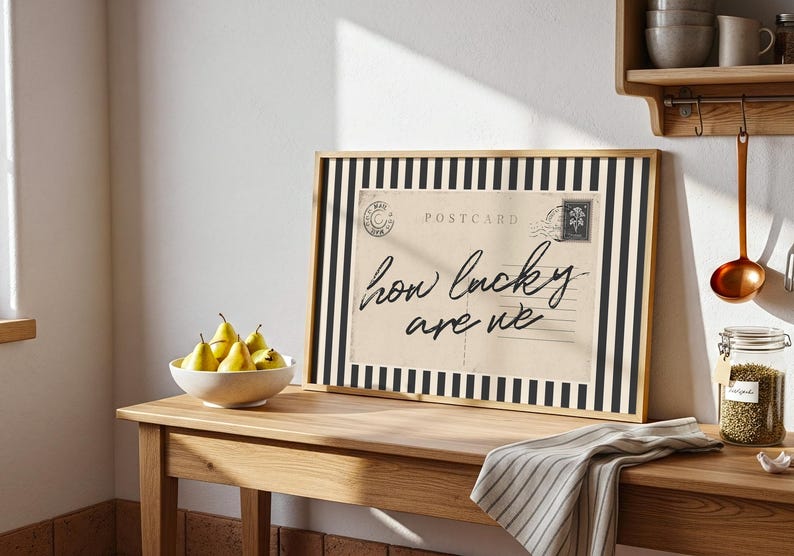 K&ouml;nnte beinhalten: Ein gerahmter Postkartendruck mit schwarz-wei&szlig; gestreiftem Rand, der den Text "how lucky are we" zeigt. Eine Schale mit gelben Birnen und ein gestreifter Stoff liegen auf einem Holztisch. Eine Kupferkelle h&auml;ngt in der N&auml;he.