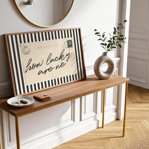 K&ouml;nnte beinhalten: Gerahmter Druck mit dem Text "how lucky are we" in Schreibschrift, vor einem Vintage-Postkartendesign mit schwarz-wei&szlig;en Streifen. Eine Holzkonsolentisch mit Vase und einer kleinen Schale mit Schl&uuml;sseln.