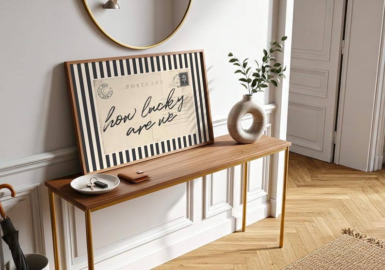 K&ouml;nnte beinhalten: Gerahmter Druck mit dem Text "how lucky are we" in Schreibschrift, vor einem Vintage-Postkartendesign mit schwarz-wei&szlig;en Streifen. Eine Holzkonsolentisch mit Vase und einer kleinen Schale mit Schl&uuml;sseln.