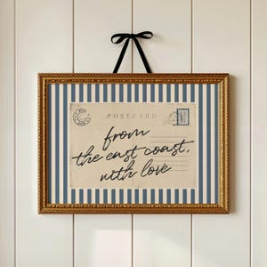 Puede incluir: Una impresión enmarcada con un marco dorado ornamentado. La impresión presenta un diseño de postal con un fondo a rayas azules y blancas. La postal dice "from the east coast, with love" en escritura cursiva. Se adjunta una cinta negra para colgar.